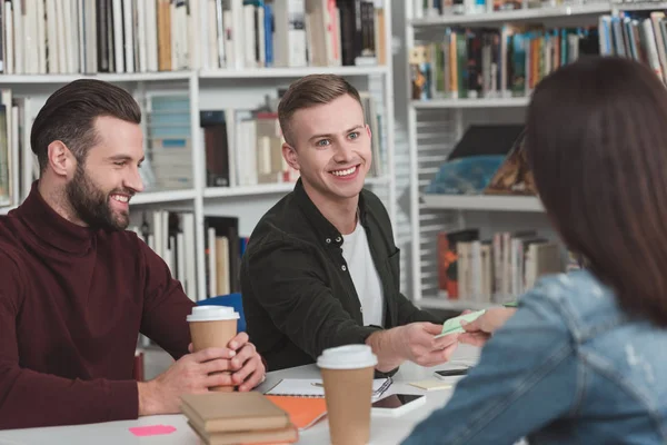 Усміхнені студенти в бібліотеці з одноразовими чашками кави — стокове фото