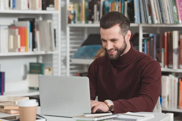Sorrindo estudante bonito usando laptop na biblioteca — Fotografia de Stock