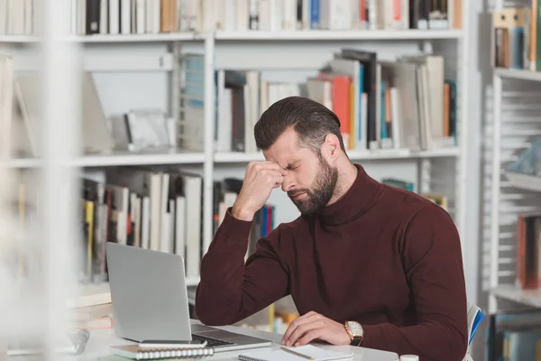 Estudante cansado tocando nariz ponte enquanto trabalhava com laptop na biblioteca — Fotografia de Stock