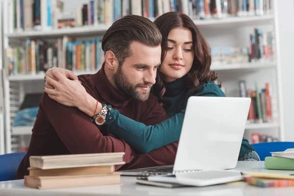 Namorada abraçando namorado e eles olhando para laptop na biblioteca — Fotografia de Stock