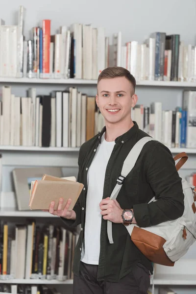 Étudiant mâle souriant avec des livres regardant la caméra dans la bibliothèque — Photo de stock