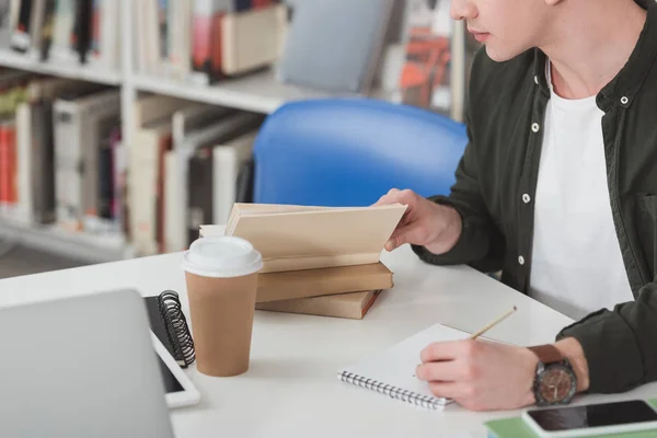 Abgeschnittenes Bild eines Schülers, der Buch liest und Notizen in der Bibliothek macht — Stockfoto