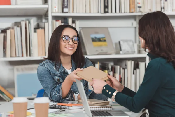 African american girl giving book to student in library — Stock Photo