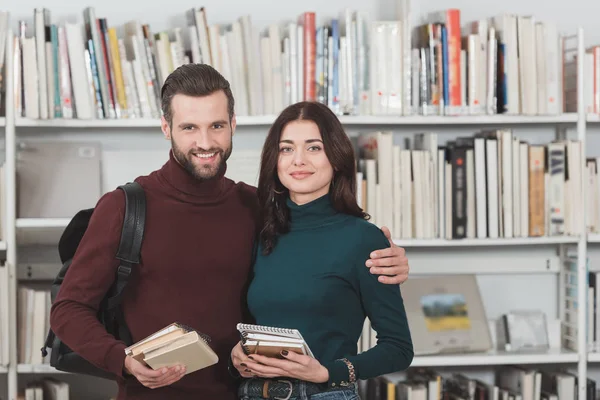 Улыбающаяся пара с книгами, смотрящими на камеру в библиотеке — стоковое фото