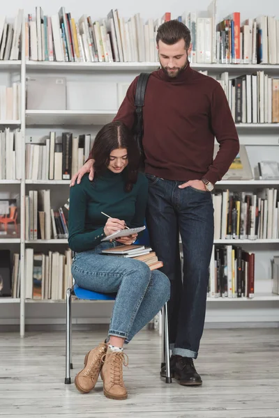Namorado de pé perto da namorada escrevendo algo para notebook na biblioteca — Fotografia de Stock