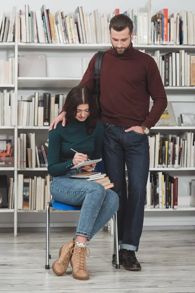 Novio de pie cerca de novia escribiendo algo a cuaderno en la biblioteca - foto de stock