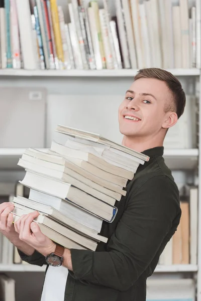 Visão lateral do estudante sorrindo segurando pilha de livros e olhando para a câmera — Fotografia de Stock