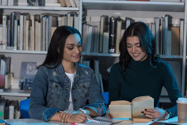 Two multicultural female friends looking at book in library — Stock Photo