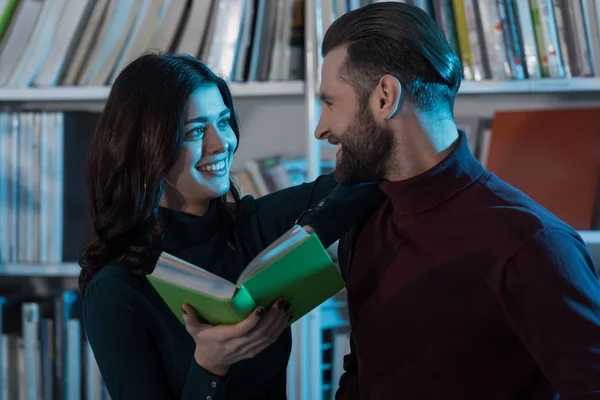 Sonriente pareja sosteniendo el libro y mirándose en la biblioteca - foto de stock