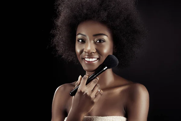 Sorridente donna afroamericana che fa trucco con pennello isolato su nero — Foto stock