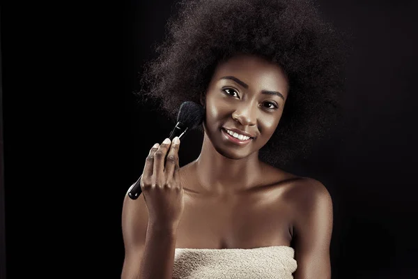 Mulher americana africana atraente fazendo maquiagem com escova isolada em preto — Fotografia de Stock