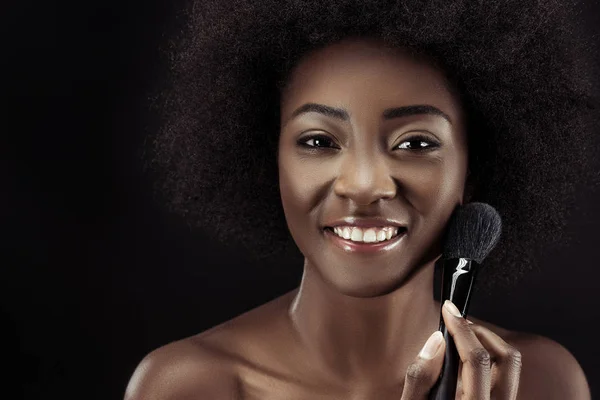 Happy african american woman doing makeup with brush isolated on black — Stock Photo