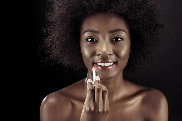 Mujer afroamericana feliz aplicando lápiz labial y mirando a la cámara aislada en negro - foto de stock