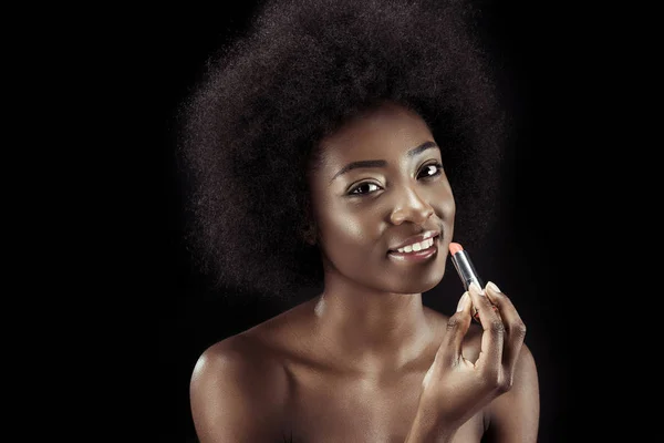 Sonriente mujer afroamericana aplicando lápiz labial y mirando a la cámara aislada en negro - foto de stock