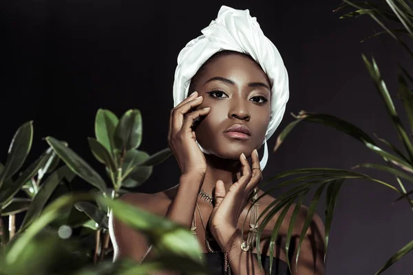 Mujer afroamericana joven de moda en elegante envoltura de cabeza de alambre detrás de las hojas - foto de stock