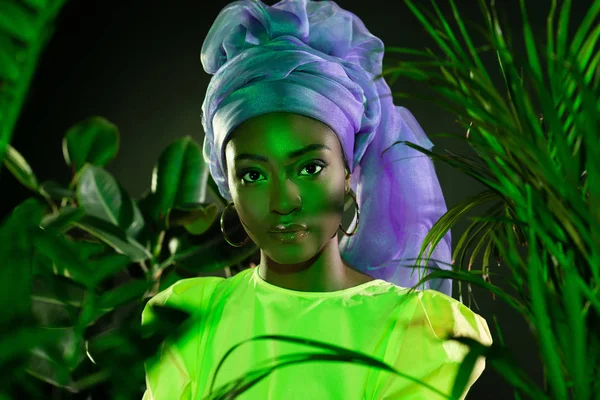 Hermosa mujer afroamericana en envoltura de cabeza de alambre tradicional bajo luz verde mirando a la cámara - foto de stock