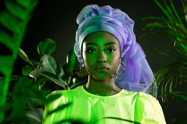 Joven afroamericana mujer en tradicional alambre cabeza abrigo bajo verde luz mirando cámara — Stock Photo