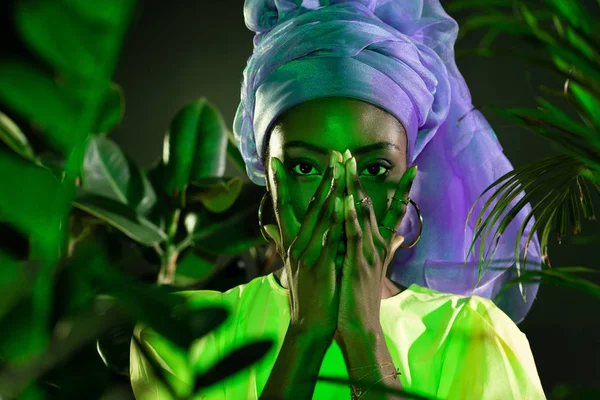 Joven afroamericana mujer en tradicional cabeza de alambre cubrir la cara con las manos bajo luz verde — Stock Photo