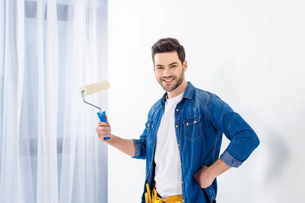 Homem sorrindo segurando pincel rolo de pintura e olhando para a câmera — Fotografia de Stock