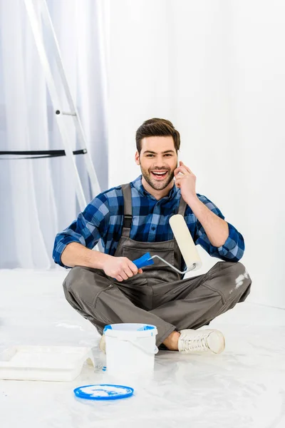 Uomo sorridente seduto con pennello rullo di vernice e parlando da smartphone — Foto stock
