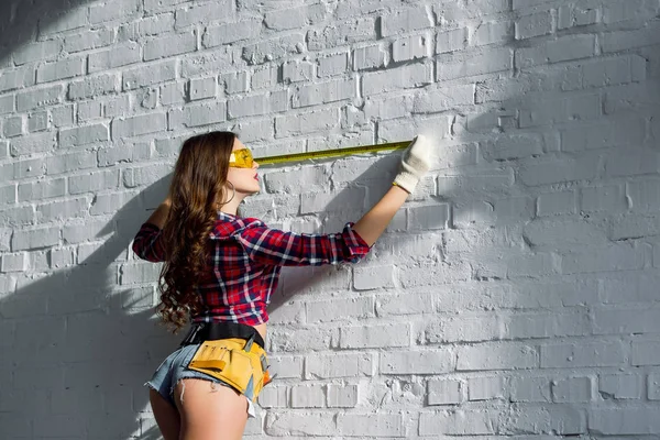 Mulher sexy com cinto ferramenta medindo parede de tijolo branco — Fotografia de Stock