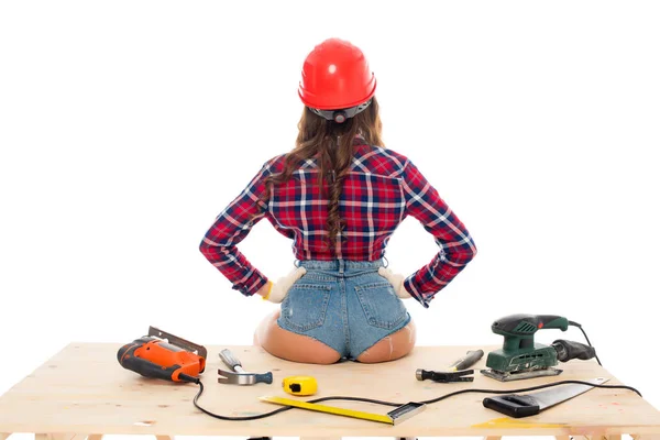 Vista trasera de chica sexy en hardhat sentado en mesa de madera con herramientas, aislado en blanco - foto de stock