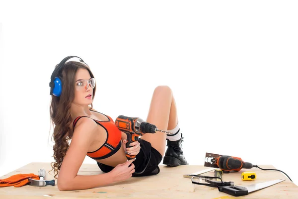 Mulher sexy em fones de ouvido de proteção posando com broca elétrica na mesa de madeira com ferramentas, isolado em branco — Fotografia de Stock