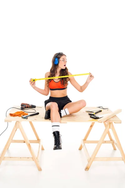 Seductive girl in protective headphones sitting on wooden table with tools, isolated on white — Stock Photo