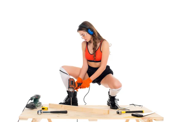 Menina bonita em fones de ouvido de proteção trabalhando com broca elétrica na mesa de madeira com ferramentas, isolado em branco — Fotografia de Stock