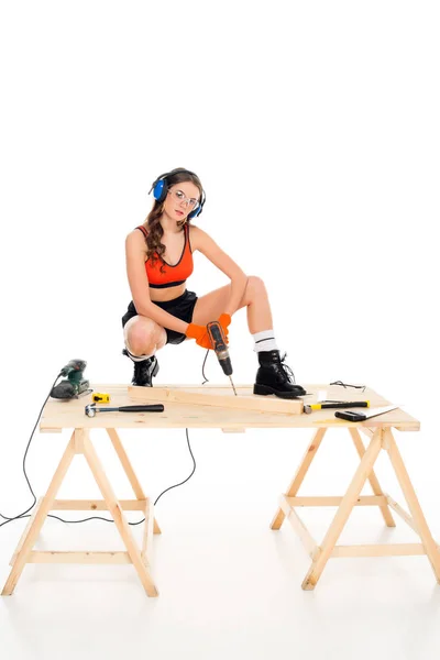 Chica en auriculares protectores trabajando con taladro eléctrico en mesa de madera con herramientas, aislado en blanco - foto de stock