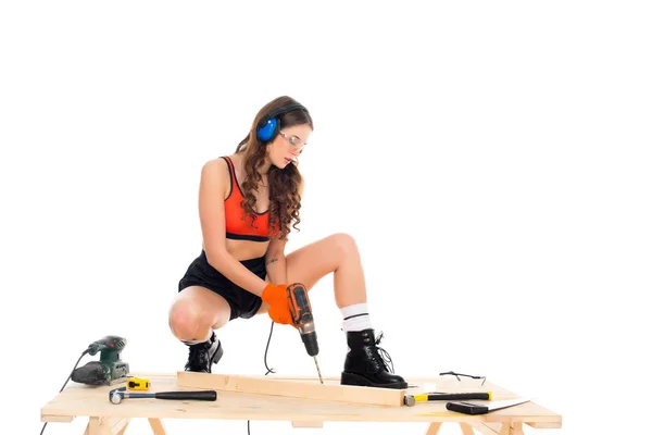 Sexy Mädchen in schützenden Kopfhörern, die mit Elektrobohrmaschine am Holztisch mit Werkzeug arbeiten, isoliert auf weiß — Stockfoto