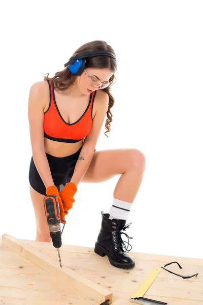 Fille sexy dans des écouteurs de protection travaillant avec perceuse électrique à la table en bois, isolé sur blanc — Photo de stock