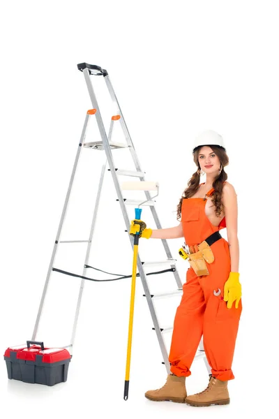 Atractiva trabajadora sosteniendo rodillo de pintura cerca de escalera y caja de herramientas, aislado en blanco - foto de stock