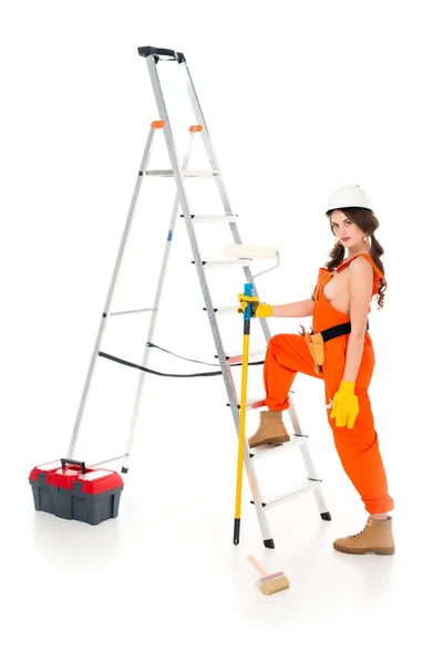 Hermosa trabajadora sosteniendo rodillo de pintura cerca de escalera y caja de herramientas, aislado en blanco - foto de stock