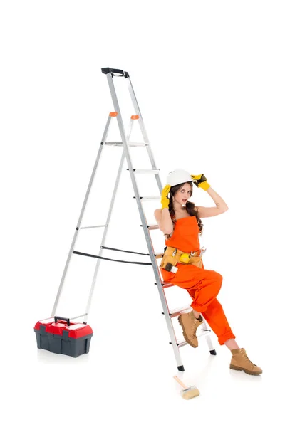 Beautiful workwoman in overalls and safety helmet near ladder and toolbox, isolated on white — Stock Photo