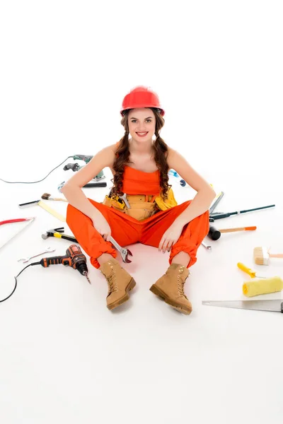 Lächelndes Mädchen in Overalls und Bollenhut, das mit verschiedenen Werkzeugen auf dem Boden sitzt, isoliert auf weißem Grund — Stockfoto