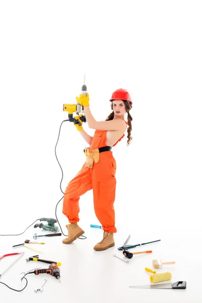 Trabajadora en overol con taladros eléctricos, diferentes herramientas tumbadas en el suelo, aisladas en blanco - foto de stock