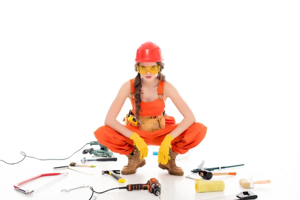 Trabajadora confiada en overoles sentada en el suelo con diferentes equipos y herramientas, aislada en blanco - foto de stock