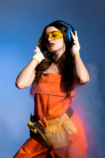 Hermosa chica en overol con cinturón de herramientas, gafas y auriculares protectores, aislado en azul - foto de stock