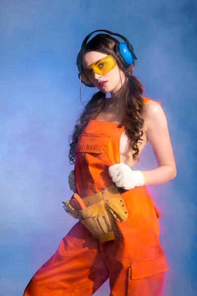 Beautiful sexy girl in overalls with tool belt, goggles and protective headphones, isolated on blue — Stock Photo