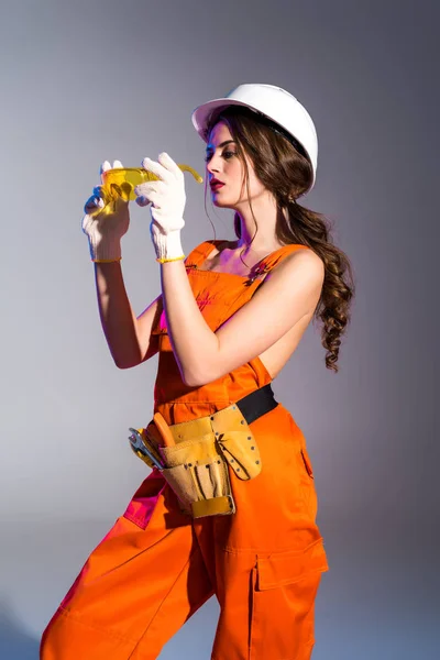 Beautiful girl in overalls with tool belt, goggles and safety helmet, isolated on grey — Stock Photo