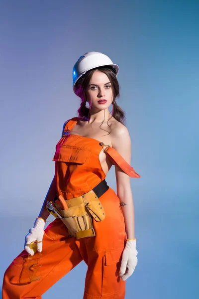 Beautiful sexy in overalls and safety helmet with tool belt and goggles, isolated on blue — Stock Photo