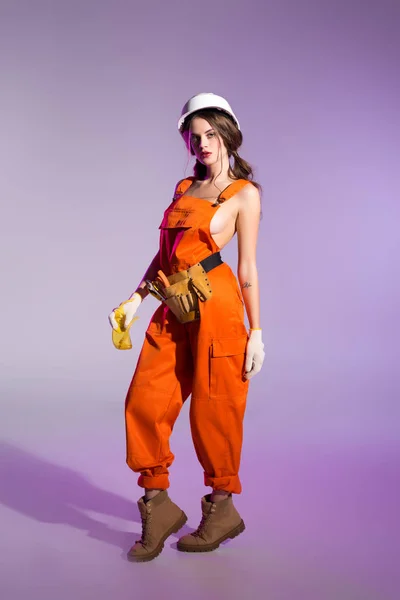 Sexy girl in uniform and safety helmet with tool belt and goggles, on purple — Stock Photo