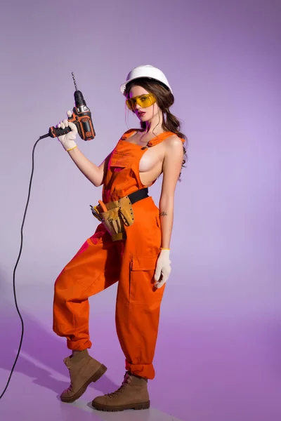 Chica atractiva en overoles y casco de seguridad con correa de herramientas que sostiene taladro eléctrico, en púrpura - foto de stock