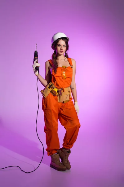 Sexy workwoman in overalls and safety helmet with tool belt holding electric drill, on purple — Stock Photo
