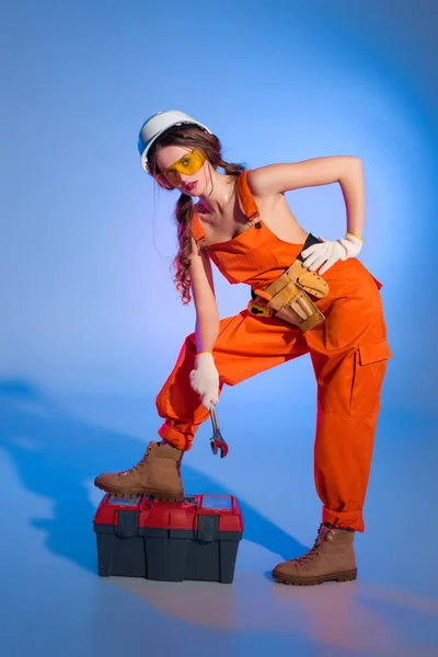 Attractive girl in overalls uniform with tool belt and toolbox, on blue — Stock Photo