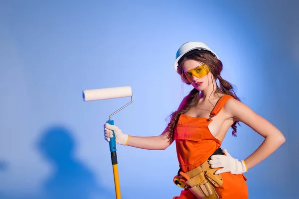 Seductive girl in overalls and safety helmet with tool belt holding painting roller, on blue — Stock Photo