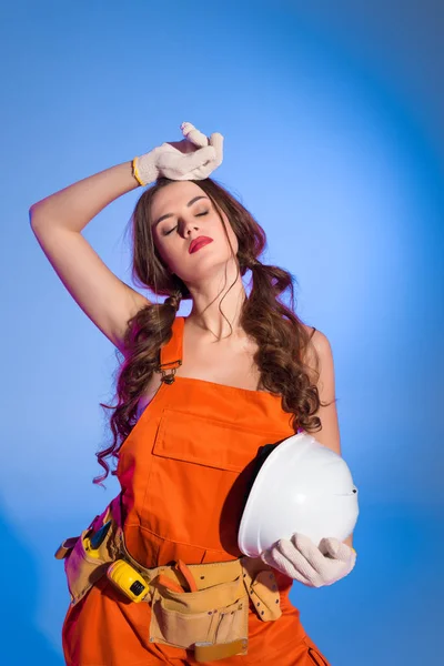 Bela menina cansada em macacão segurando capacete de segurança, isolado em azul — Fotografia de Stock