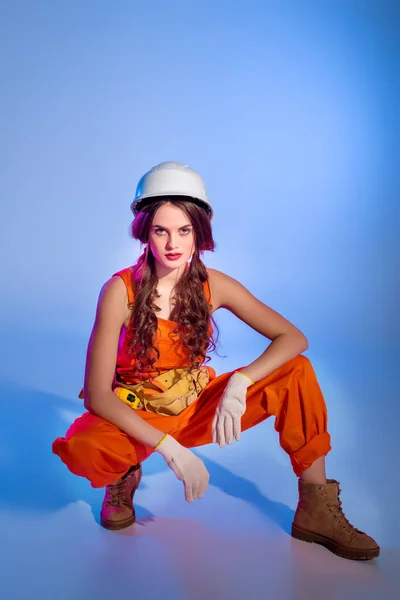 Beautiful girl in overalls and safety helmet, on blue — Stock Photo