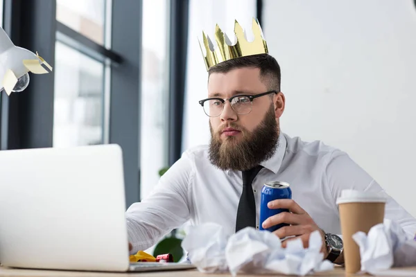Портрет бізнесмена з паперовою короною на голові і содовим напоєм в руці, що працює на ноутбуці на робочому місці в офісі — стокове фото